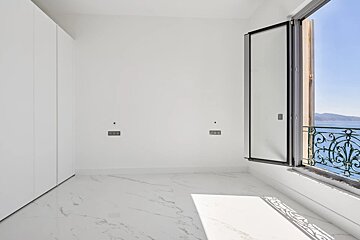 Empty white room with marble floor and built-in wardrobe. A large window with an ornate railing offers a view of the sea and distant mountains.