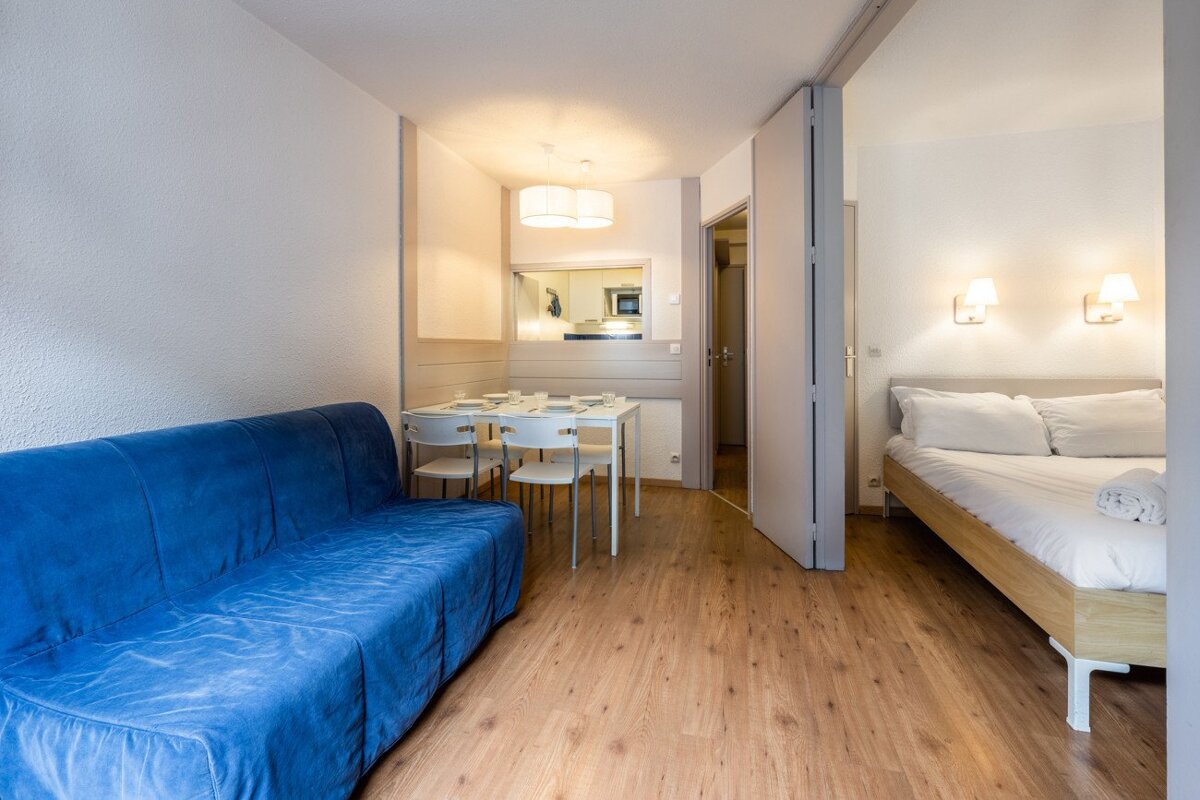 A modern studio apartment featuring a blue sofa, dining table, visible kitchenette, and a bedroom area with a bed and wooden floor.