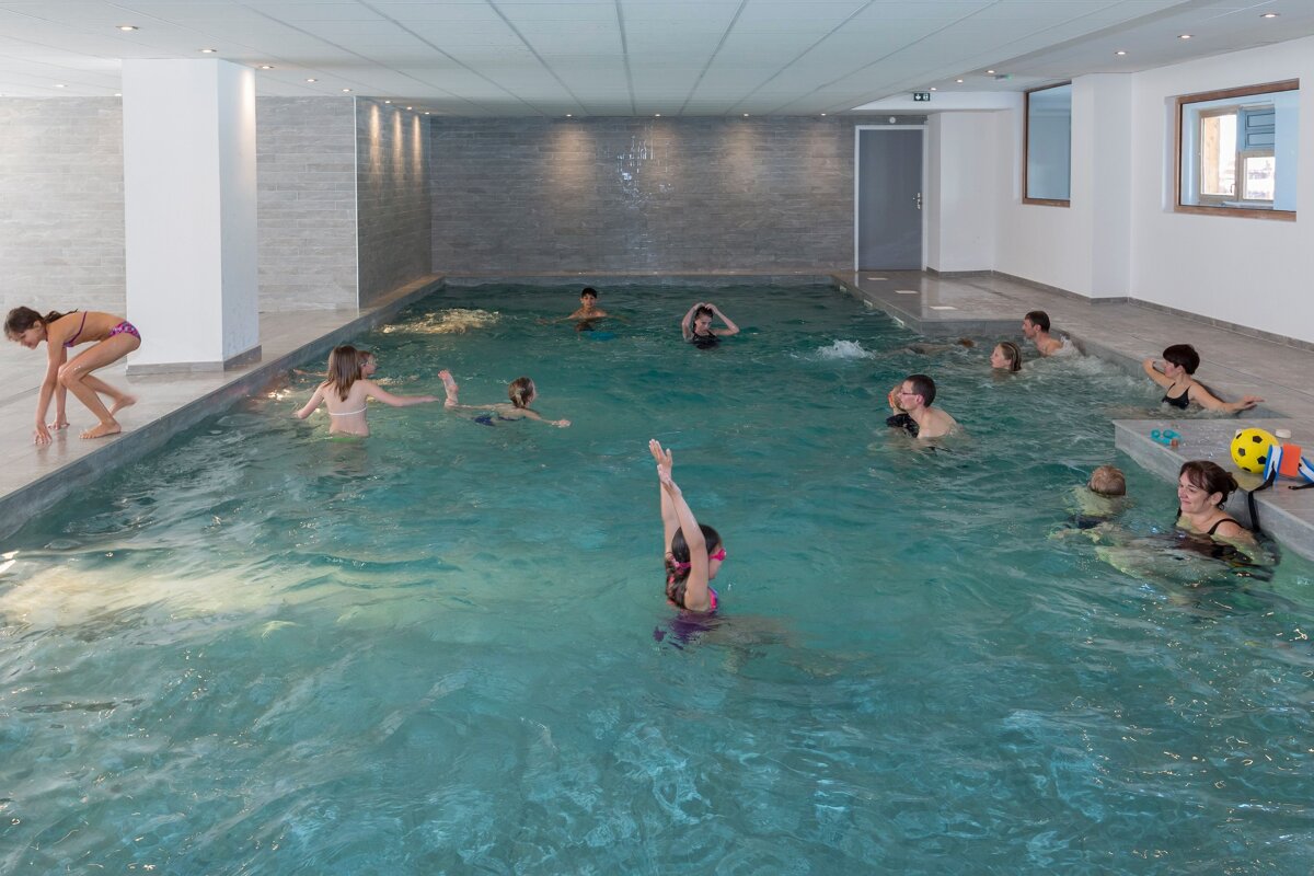 An indoor swimming pool with a mix of adults and children swimming, playing, and relaxing. Some are in the shallow end, others are swimming.