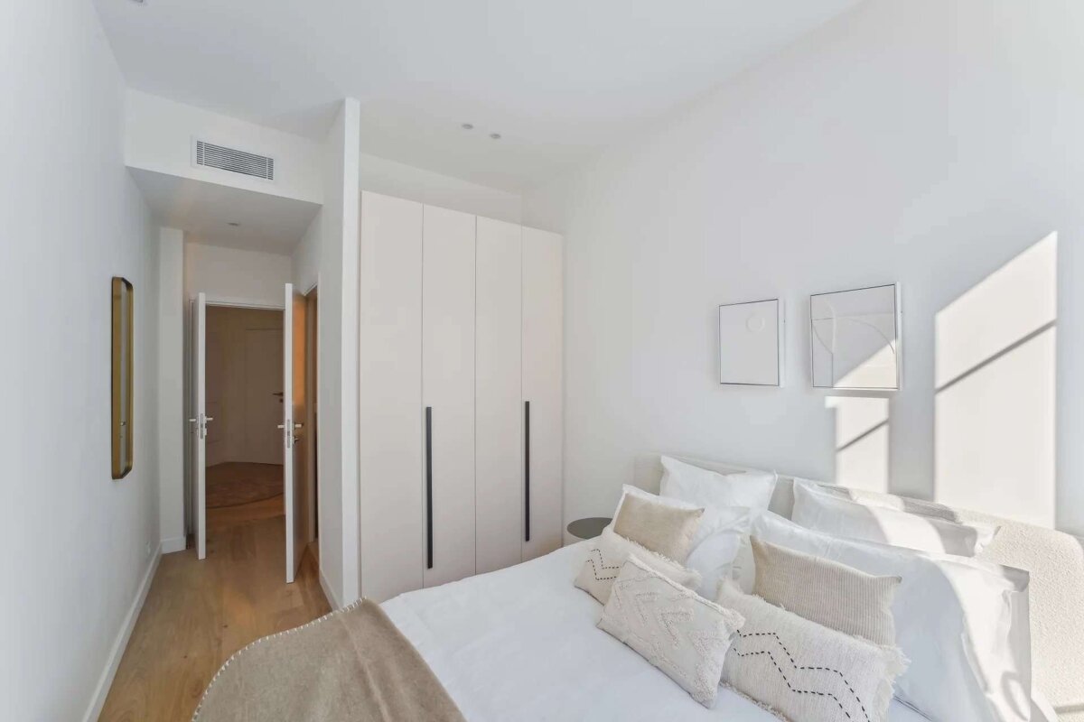 A minimalist white bedroom with a bed, textured pillows, built-in wardrobe, and a sunlit wall. A hallway with open doors is visible.