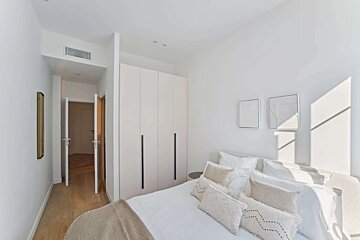 A minimalist white bedroom with a bed, textured pillows, built-in wardrobe, and a sunlit wall. A hallway with open doors is visible.