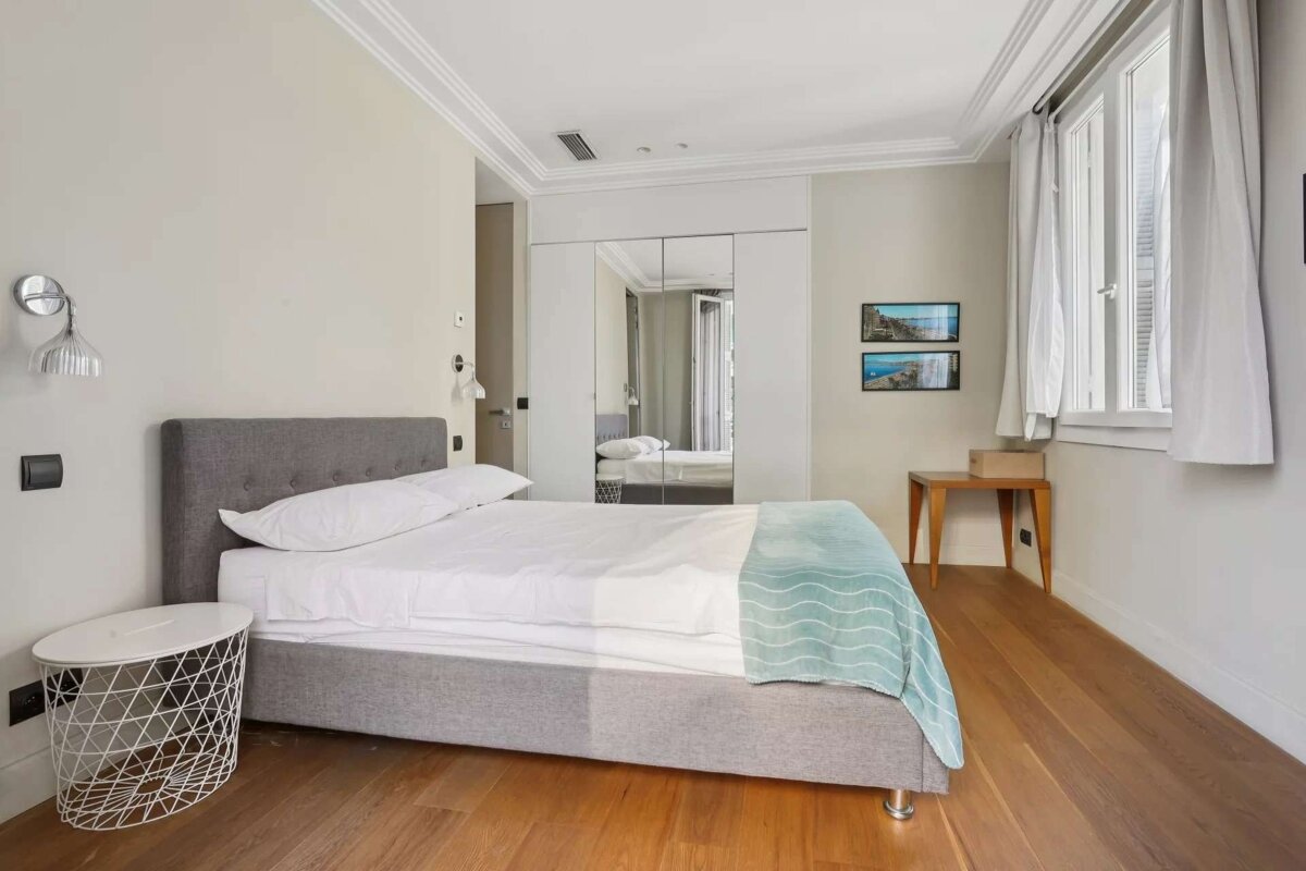 A modern bedroom features a grey upholstered bed with white bedding & a teal throw, hardwood floors, mirrored closet, window, and wall art.