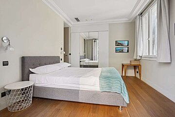 A modern bedroom features a grey upholstered bed with white bedding & a teal throw, hardwood floors, mirrored closet, window, and wall art.