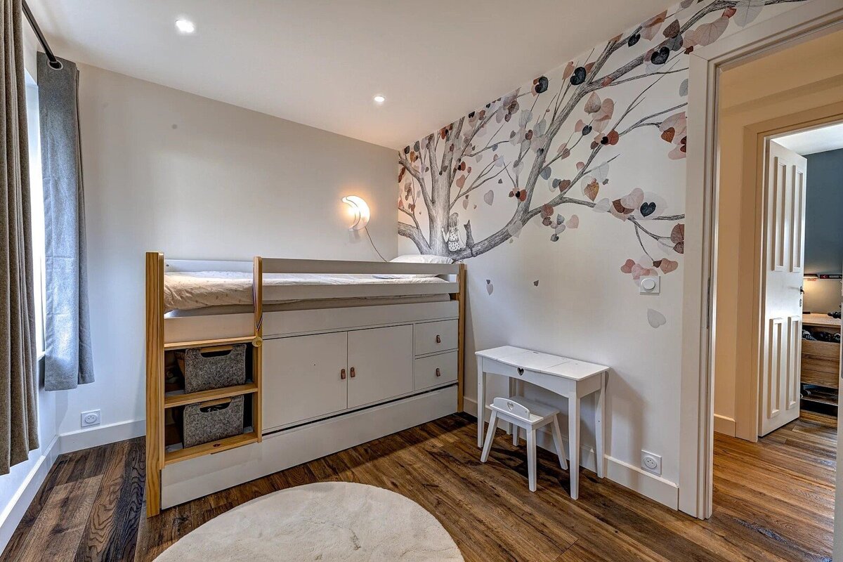 Bright kid's room with a loft bed & storage, whimsical tree mural, small desk, and warm wooden floor with a white rug.