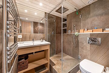 A modern bathroom with brown tiled walls, a glass shower stall, wooden vanity, toilet, and heated towel rack.