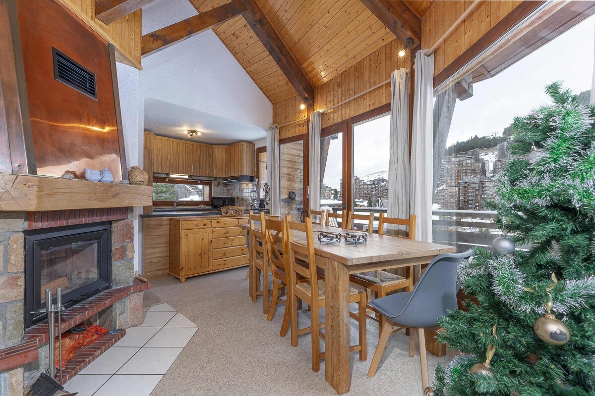 Cozy ski chalet interior with a fireplace, wooden dining table, kitchen, and large windows offering a snowy mountain view, plus a Christmas tree.