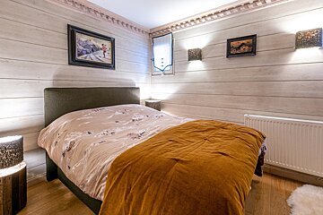 A cozy bedroom with light wood-paneled walls, a double bed, orange throw, and ski-themed art. Warm lighting and wooden floors create a rustic feel.