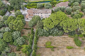 Aerial view of a large, traditional house with a red-tiled roof, nestled among abundant green trees and terraced, dry land.
