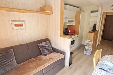 A cozy, compact living space with light wood-paneled walls. It features a grey sofa, an open-plan kitchenette with white cabinets, and a dining area.