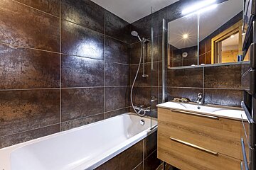 A modern bathroom with a white tub/shower, dark metallic-look tiled walls, and a light wood vanity with a mirror.