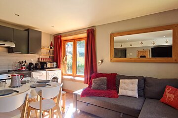 A bright, open-plan studio apartment. Features a modern kitchen, dining area, and living space with a grey sofa, red accents, and a large mirror.