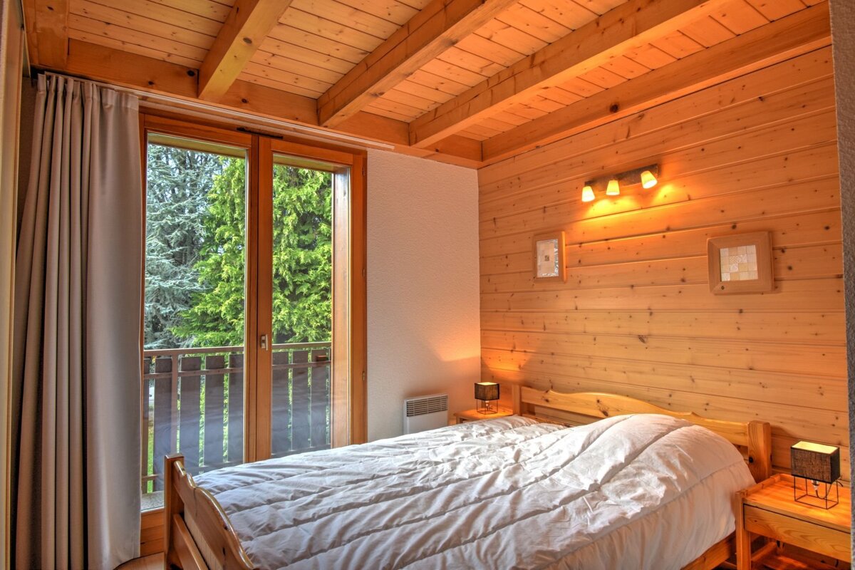 A cozy wooden bedroom with wood-paneled walls and ceiling, a bed with white covers, and a large window opening to a lush green forest view.