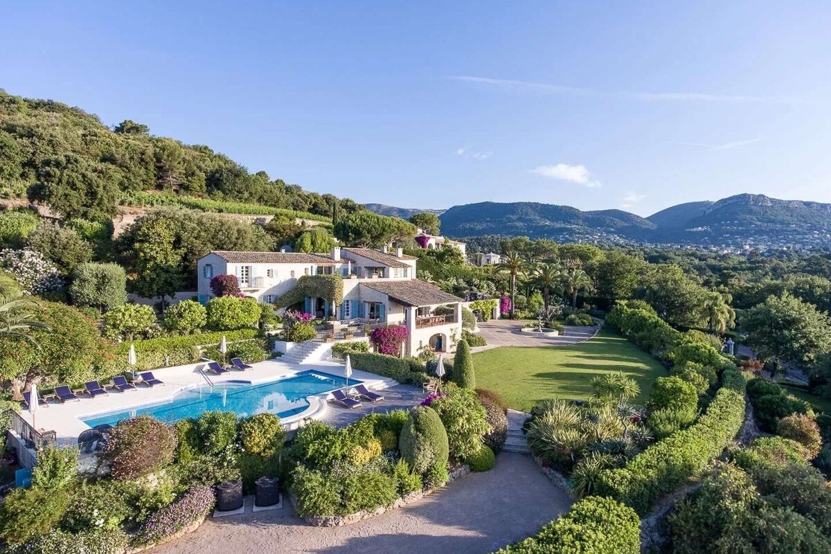 An aerial view of a sprawling villa with a large swimming pool, extensive manicured gardens, and distant mountains under a clear sky.