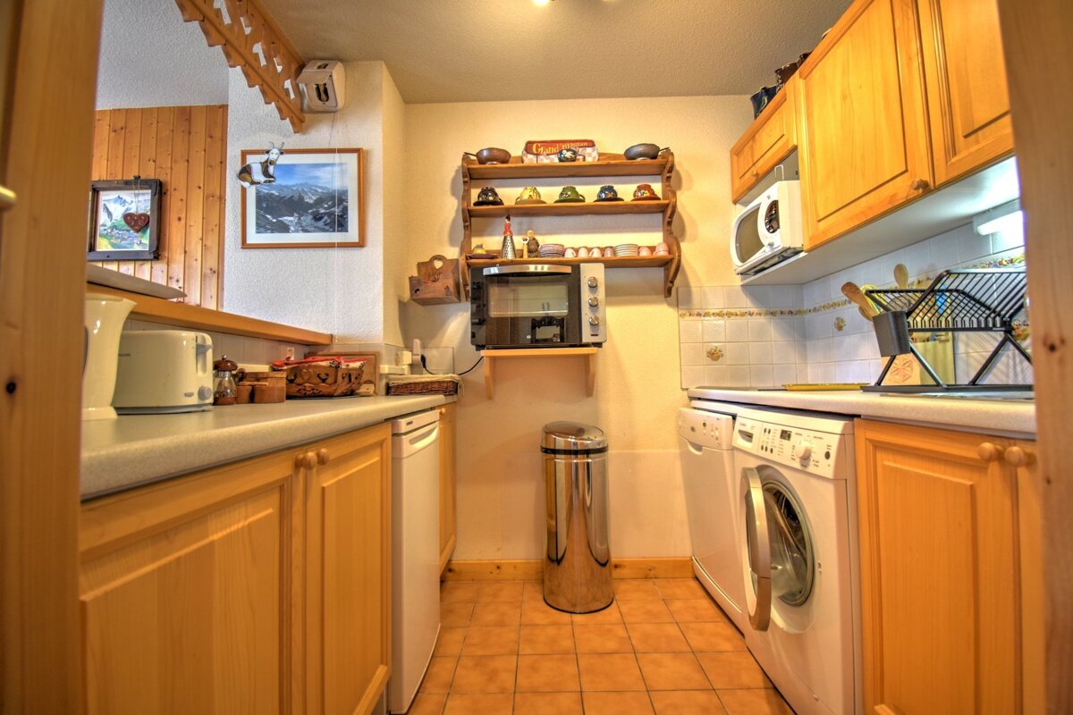 A narrow kitchen with wooden cabinets, tile floors, a washing machine, dishwasher, and small appliances.