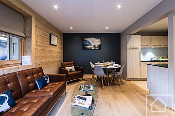 Modern chalet living room featuring leather sofas, wood-paneled walls, a glass coffee table, a dining set, and an open kitchen.
