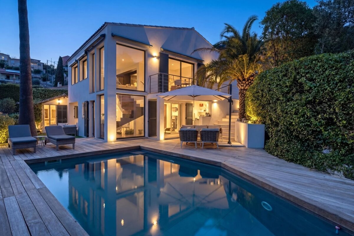 A modern, illuminated villa with a swimming pool and wooden deck at dusk.