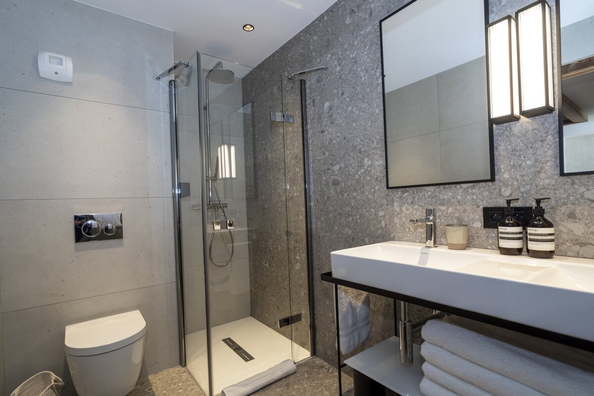 A modern gray bathroom with a glass walk-in shower, wall-mounted toilet, and a sleek vanity with sink, mirror, and stacked white towels.