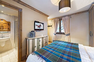 Cozy bedroom with a plaid bed, wall-mounted TV, dresser, window, and an en-suite bathroom with a sink and shower.
