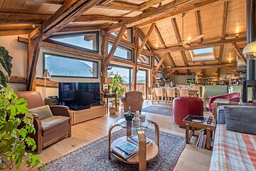 Spacious, rustic-modern open-plan living area with exposed wood beams, large windows showcasing mountain views, and integrated dining/kitchen spaces.