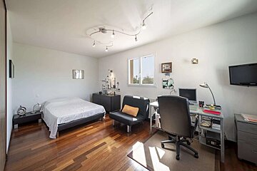 A modern bedroom featuring a bed, a workstation with a desk, computer, and office chair, a TV, and a window, all on a wooden floor.