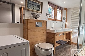 A rustic bathroom featuring a wooden toilet enclosure, double sink vanity, washing machine, and shower. A painting hangs above the toilet.