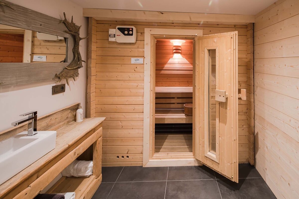 Rustic wooden bathroom with a modern sink, antler mirror, and a brightly lit sauna with its door ajar.