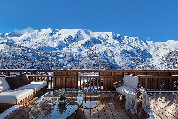 A luxurious wooden balcony with outdoor furniture overlooks a stunning, snow-covered mountain under a clear blue sky.
