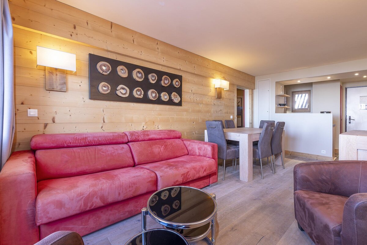 A warm living and dining room with wooden walls, a vibrant red sofa, a dining table with chairs, and contemporary art.
