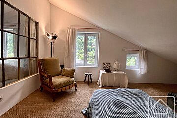 A cozy attic bedroom with a sloped ceiling, jute rug, and a rustic armchair. Windows reveal lush greenery, illuminating the simply decorated space.