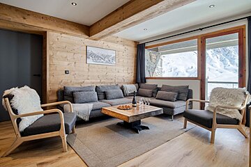 Alpine cabin living room featuring wooden paneling, grey seating with faux fur throws, and a large window overlooking snowy mountains.