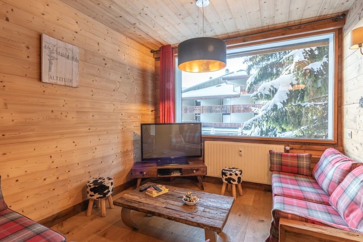 Cozy wooden cabin living room with plaid sofas, a TV, and a large window looking out onto a snow-covered pine tree.