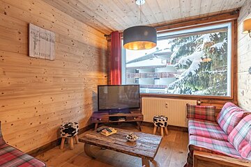 Cozy wooden cabin living room with plaid sofas, a TV, and a large window looking out onto a snow-covered pine tree.