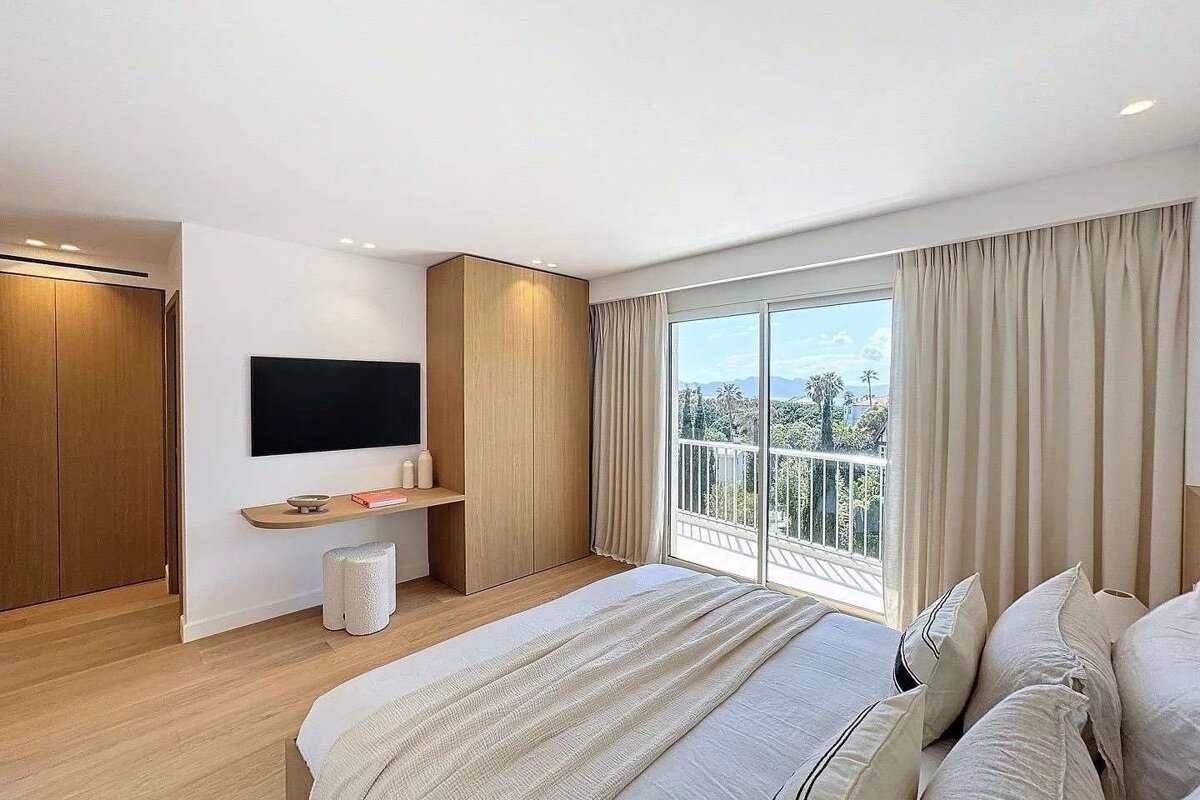 A modern, bright bedroom features a large bed, wood cabinetry, TV, and floor-to-ceiling glass doors leading to a balcony with a scenic view.