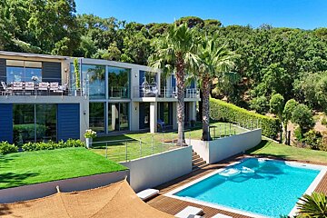 Modern multi-story villa with glass facades and balconies, a blue swimming pool, palm trees, and lush green hills in the background under a clear sky.