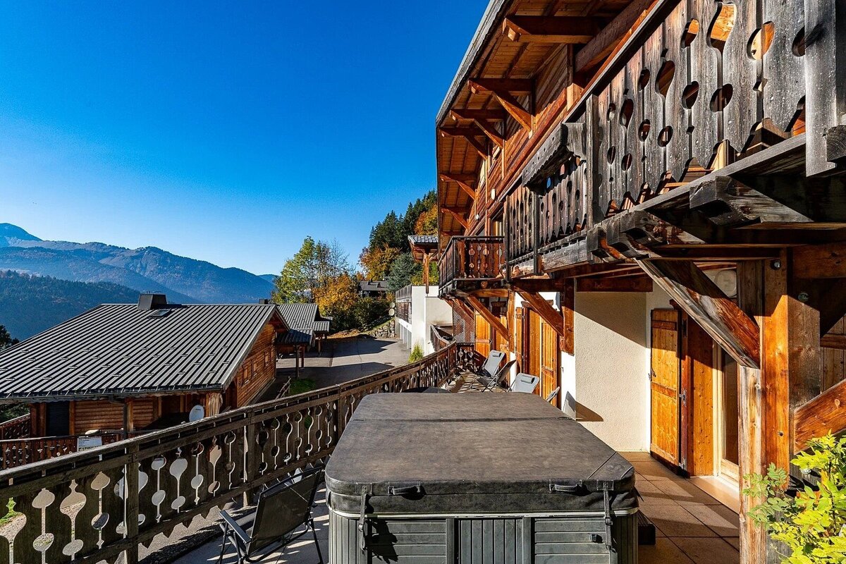 A wooden mountain chalet balcony with a covered hot tub offers panoramic views of mountains and other chalets under a clear blue sky.