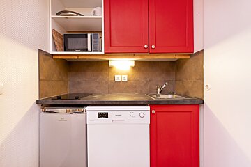 A small, modern kitchen features red upper and lower cabinets, a white dishwasher, compact fridge, microwave, sink, and cooktop. Tiled backsplash.