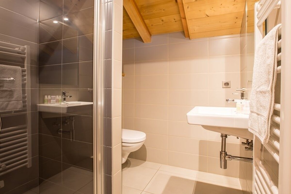 A modern tiled bathroom featuring a white sink and toilet under a wooden ceiling, with a glass shower enclosure visible on the left.