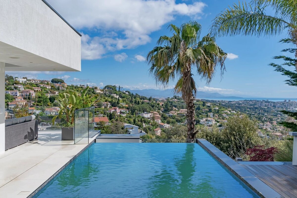 An infinity pool at a modern villa overlooking a scenic hillside town with palm trees and the distant sea under a bright blue sky.