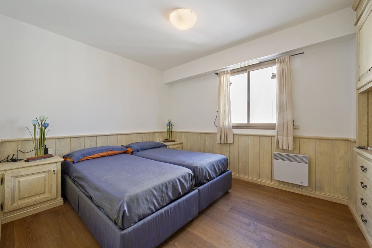 A bedroom with two twin beds, blue bedding, white and wood-paneled walls, hardwood floor, a window with curtains, and a nightstand.