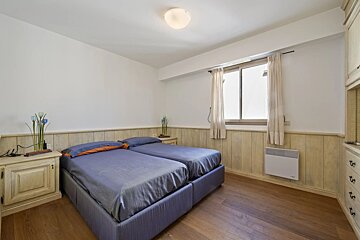 A bedroom with wooden floors, light-colored walls, two single beds with blue sheets, a window, and wooden furniture.