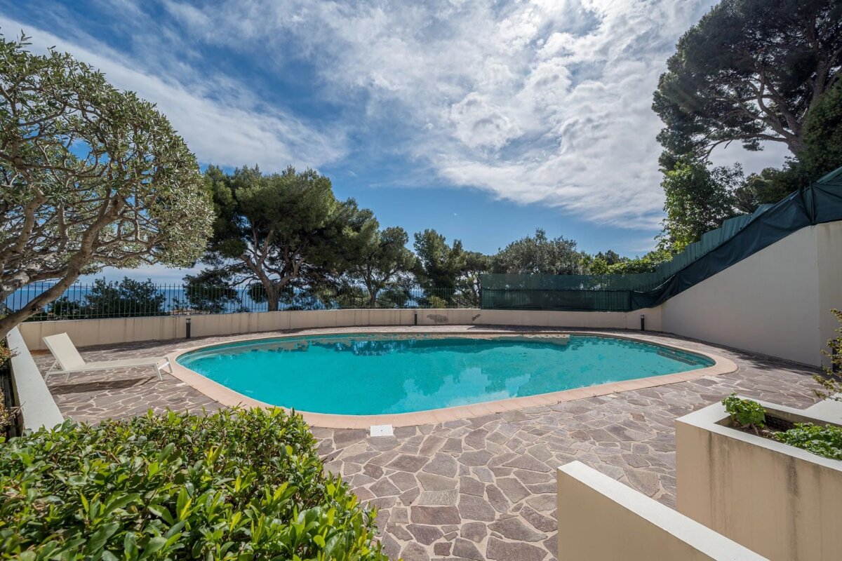 An outdoor swimming pool with blue water, surrounded by a stone patio and lush green trees under a partly cloudy blue sky. A lounge chair sits nearby.