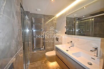 Modern bathroom featuring a double sink vanity, a large illuminated mirror, a toilet, chrome towel warmer, and sleek marble-style tiling.