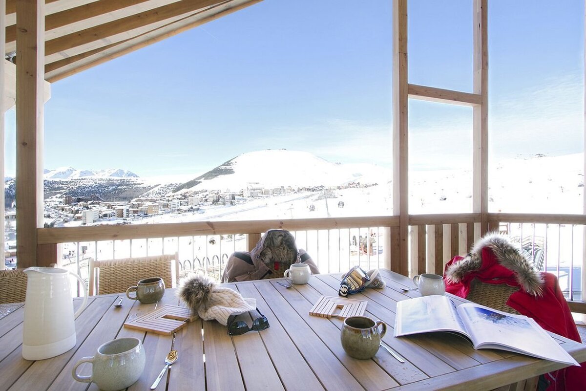 A table on a sunlit balcony with mugs and ski gear overlooks a snowy mountain resort and vast white peaks on a clear winter day.