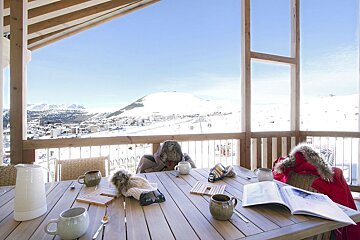 A table on a sunlit balcony with mugs and ski gear overlooks a snowy mountain resort and vast white peaks on a clear winter day.