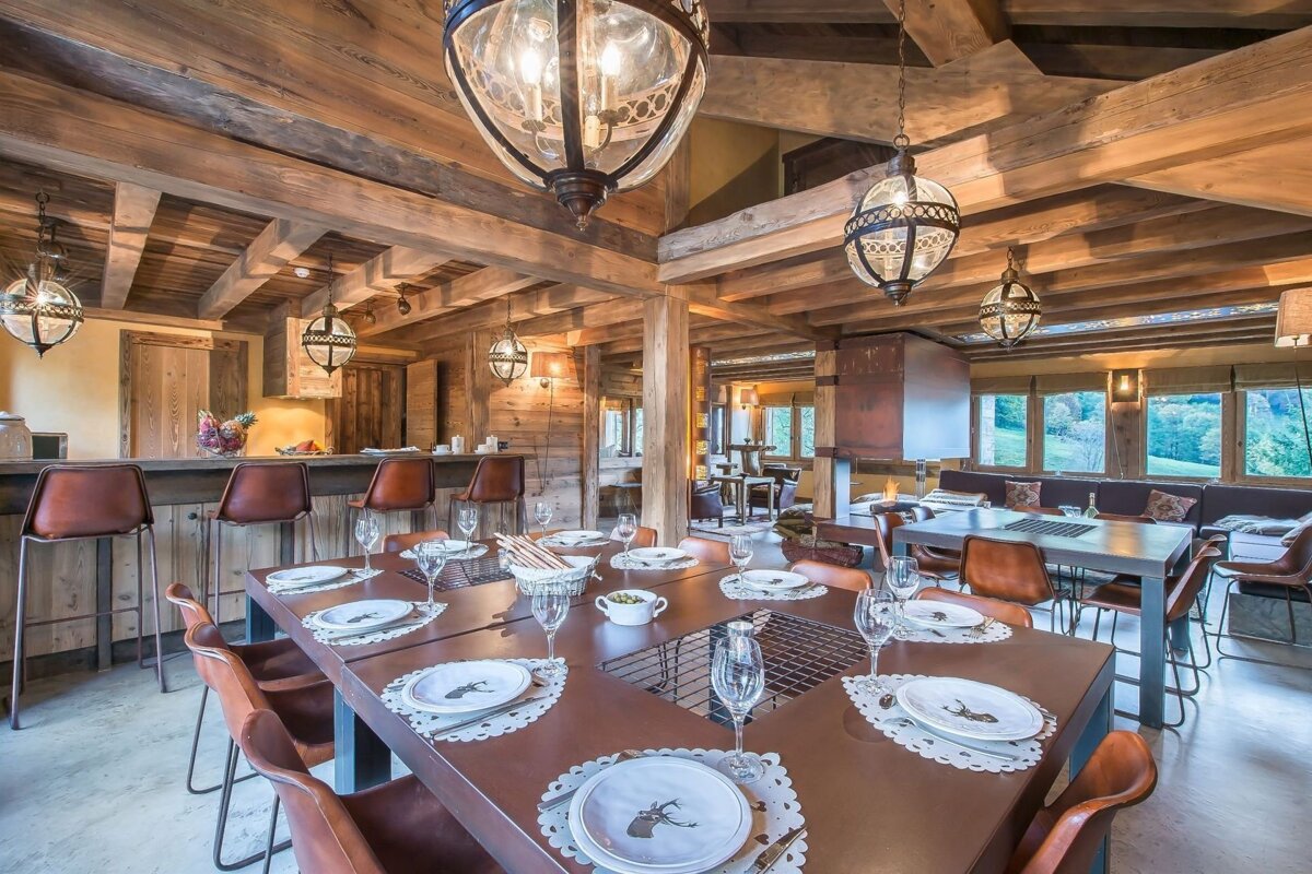 A warm, rustic chalet interior featuring wooden beams, ornate hanging lights, a large dining table, a bar, and a lounge area with outdoor views.
