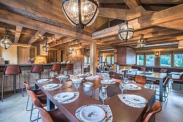 A warm, rustic chalet interior featuring wooden beams, ornate hanging lights, a large dining table, a bar, and a lounge area with outdoor views.