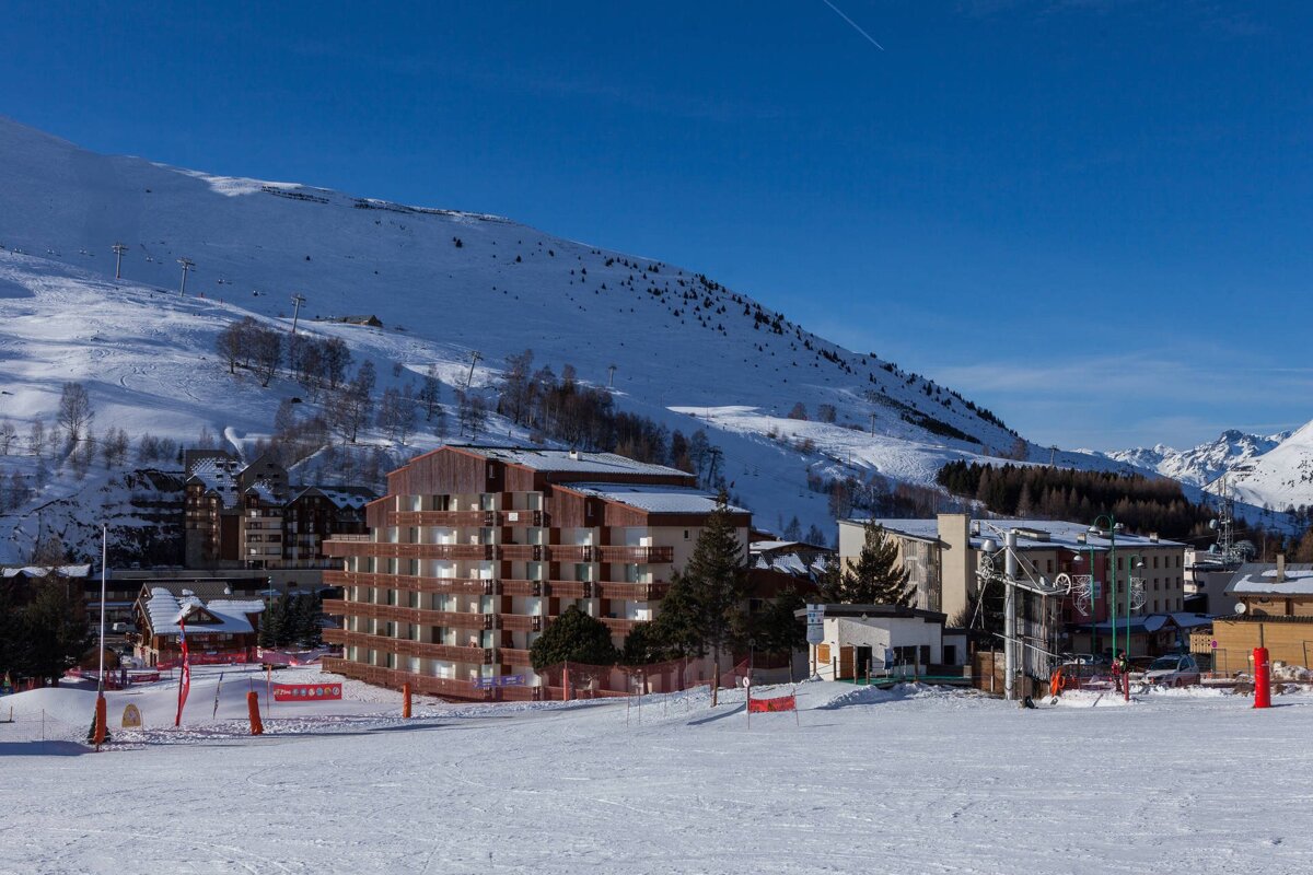 A snow-covered ski resort with buildings and slopes, surrounded by mountains under a bright blue sky.