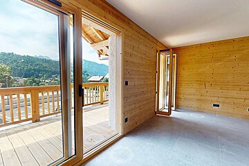 An empty room with wooden walls and light floor features large glass doors leading to a wooden deck with a stunning mountain and village view.