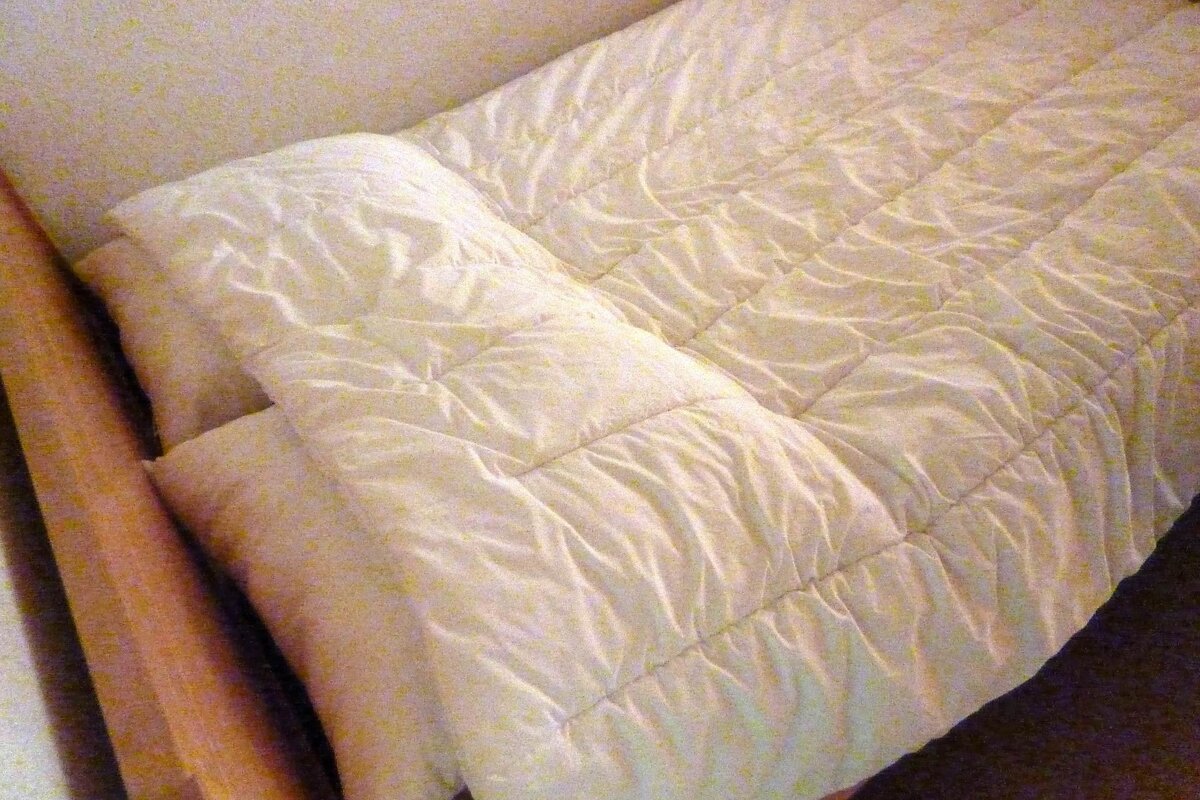 A bed with a cream-colored, quilted comforter and pillow. Part of a wooden bed frame is visible on the left side.