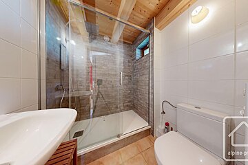 A modern bathroom features a glass-enclosed shower, white sink, toilet, white tiled walls, brick-patterned shower walls, and a wooden beam ceiling.